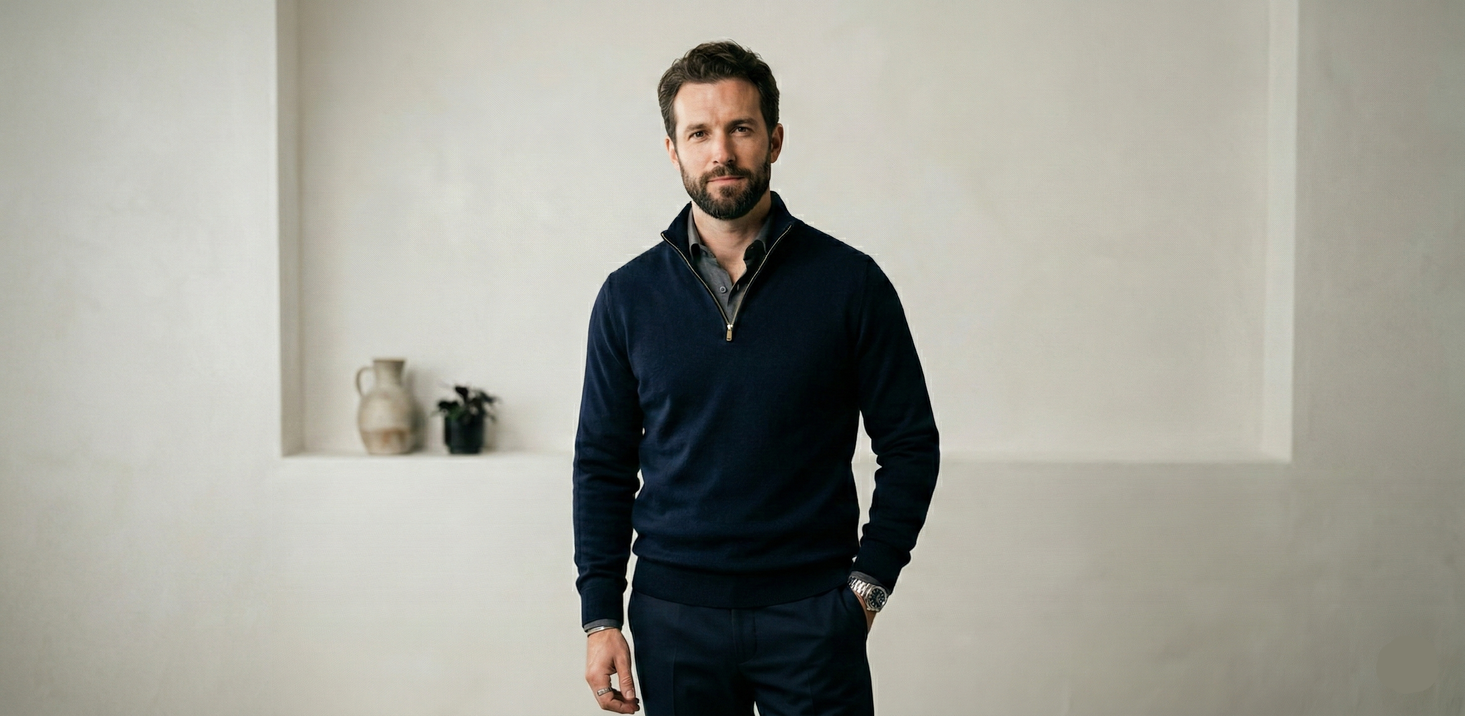 Man wearing a dark blue sweater and pants standing in a minimalistic room.